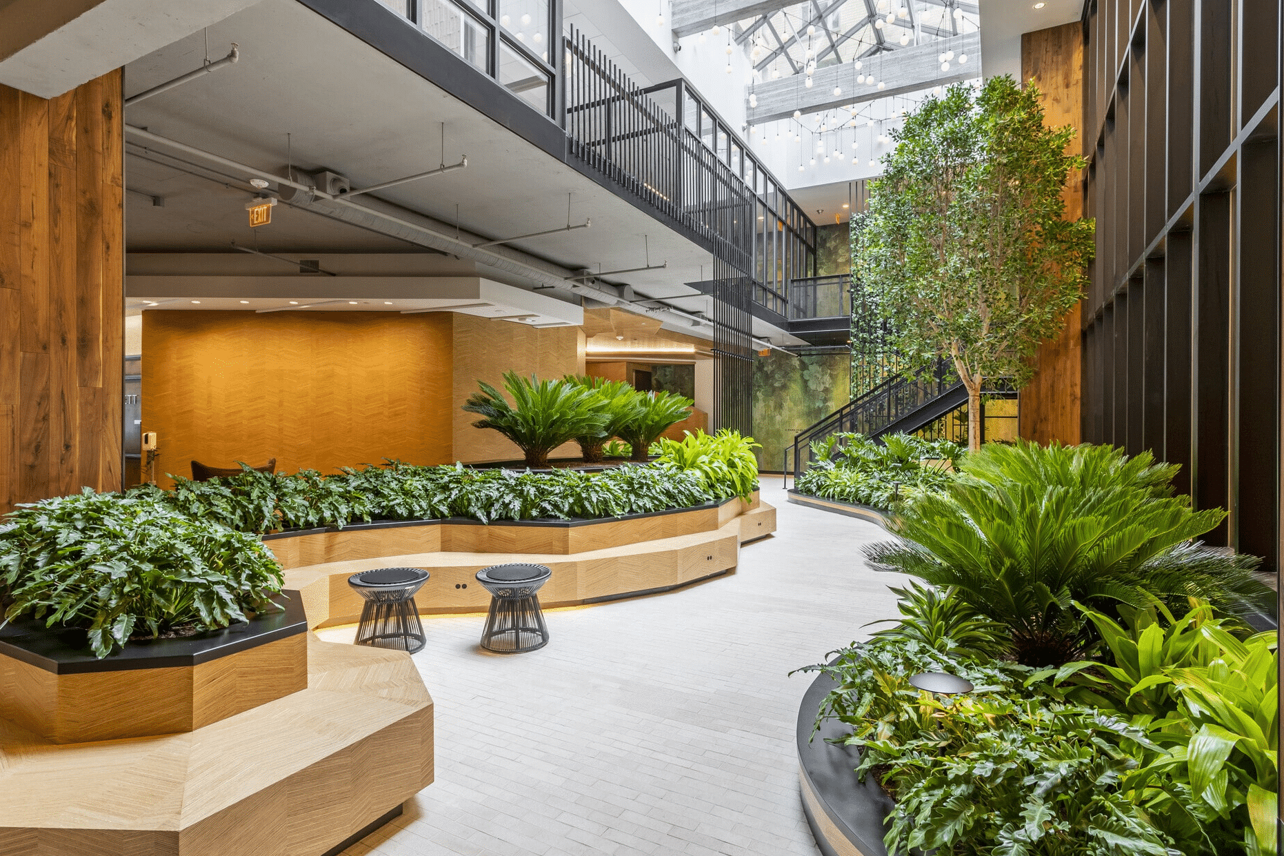 Indoor atrium with wooden planters, green plants, two small black stools, and a staircase leading to an upper level under a skylight ceiling.